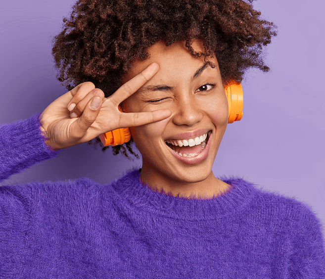 Fröhliche junge Frau mit Kopfhörern, die eine Peace-Geste macht, lächelnd, energisch und modisch, vor einem lilafarbenen Hintergrund.