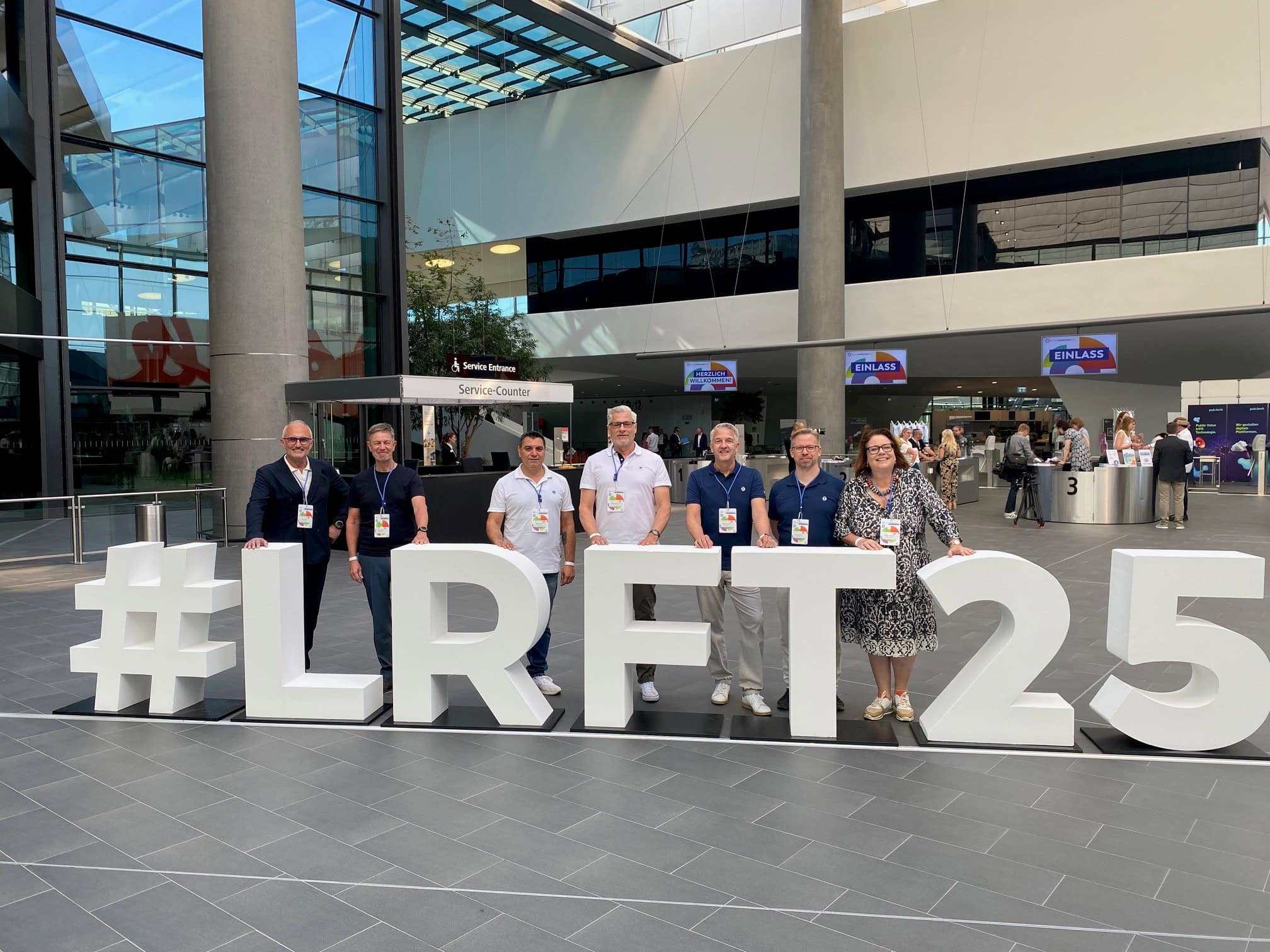 Team in einem modernen Messehalle-Design in Deutschland, mit großen #LRT25 Schild und mehreren Teilnehmern, die die Zukunft der Kreativität feiern.