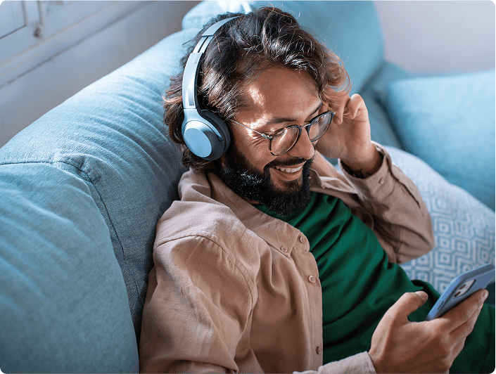 Entspannter Mann mit Kopfhörern, der auf seiner Couch Musik oder Podcasts hört und auf seinem Smartphone liest.